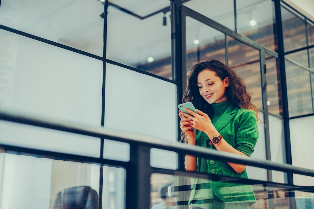 Woman in green blazer outside of an office building smiling at her mobile device that is safely managed with MDM